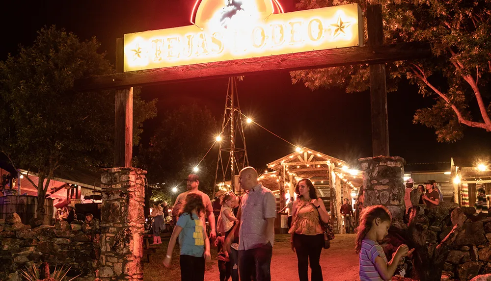 People are walking under a brightly lit sign for Tejas Rodeo at night