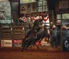 A rodeo rider is mounted on a bucking bull inside an arena surrounded by spectators and rodeo personnel