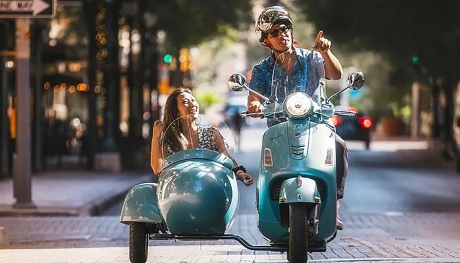 Two people are enjoying a sunny day ride on a retro blue scooter with a sidecar down an urban street.