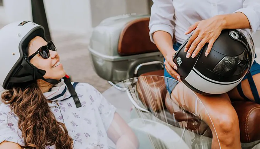 A person wearing a white helmet is smiling while another individual is seated beside them with a black helmet on their lap.