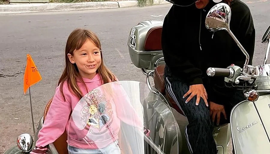 A young girl smiles while seated on a classic Vespa scooter with a clear windshield, next to an adult wearing a black sweatshirt.