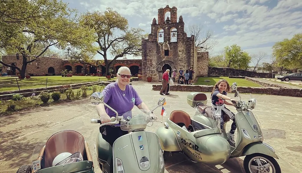 Two people are happily posing on Vespa scooters with sidecars in front of a historic stone mission building under a bright blue sky