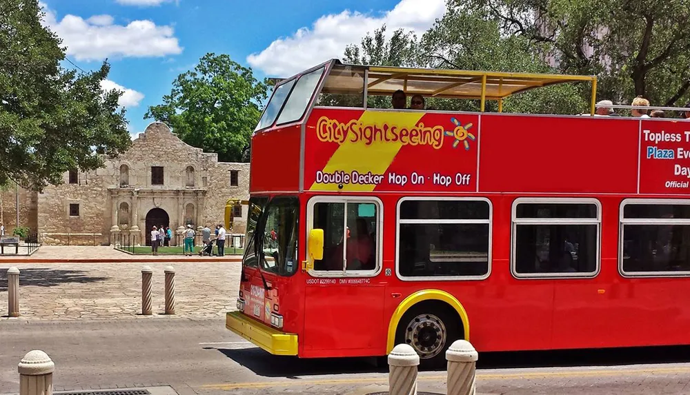 A red double-decker sightseeing bus passes by a historical stone mission building under a clear sky