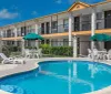 This image shows a sunny outdoor scene with a clear blue swimming pool in the foreground and a two-story motel with balcony access rooms in the background complete with umbrellas and seating arrangements around the pool area