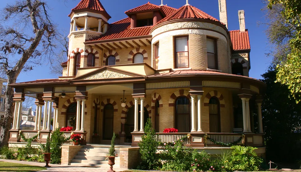 The image shows a large ornate two-story Victorian-style house with a distinctive tower arched windows a wrap-around porch with columns and well-maintained landscaping under a clear blue sky