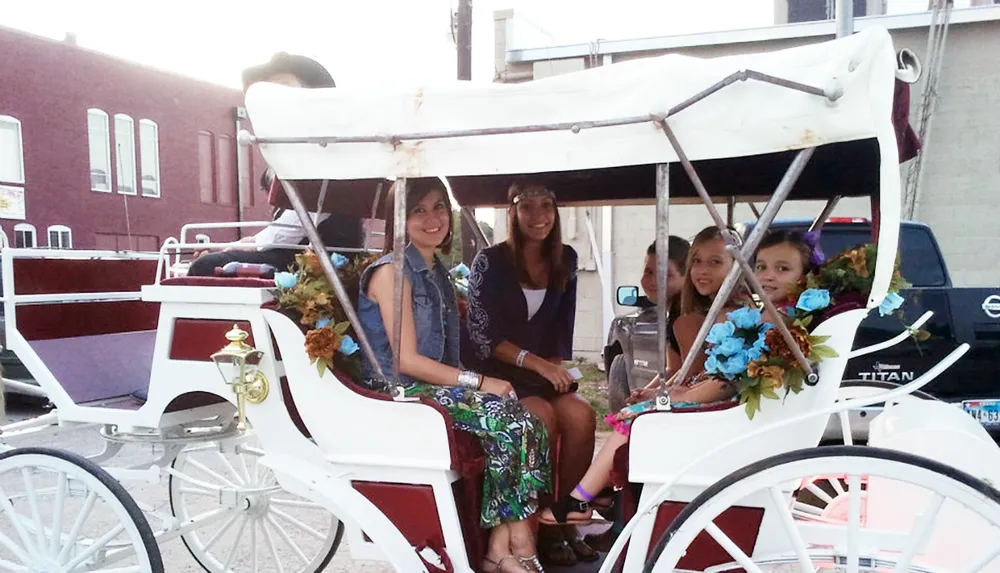 Four individuals are smiling and posing for a photo while seated in a decorative horse-drawn carriage on a city street