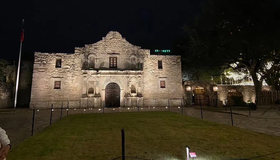 The image shows the Alamo Mission illuminated at night with a large Texas flag flying on the left and a neon sign above the right side of the complex.