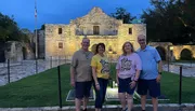 Four individuals are posing for a photo in the evening in front of the historic Alamo mission building.