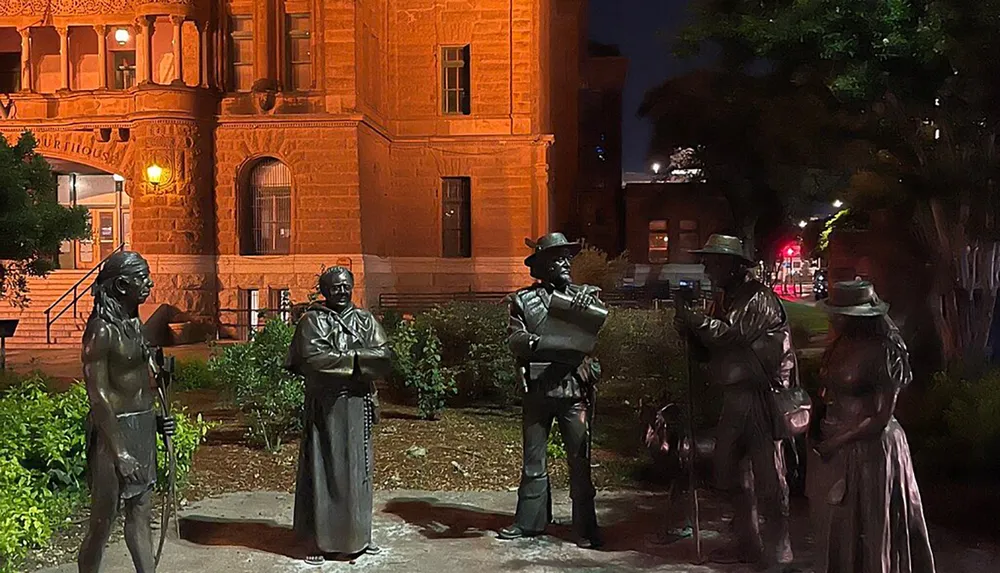 The image shows a nighttime scene featuring a group of life-sized bronze statues depicting historical figures in front of a lit-up courthouse building