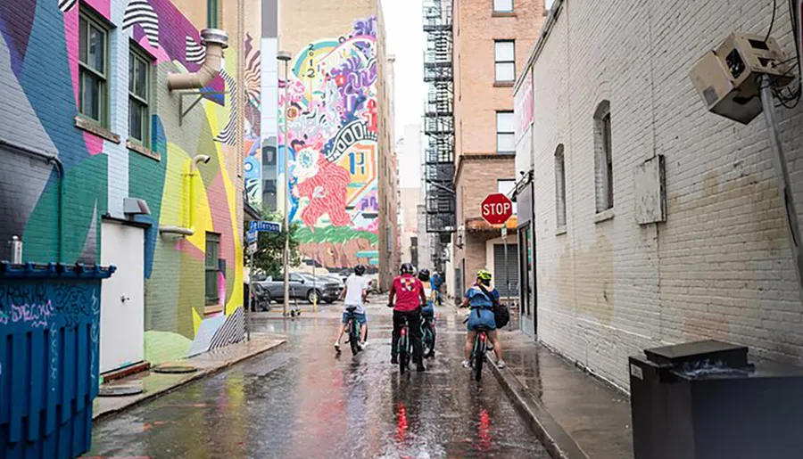 Cyclists ride through an urban alley adorned with vibrant street art after a rain.