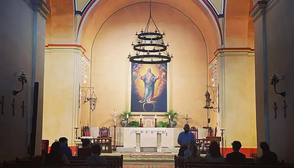 This image depicts the interior of a church with people seated in pews focusing on an altar with a large painting of Jesus Christ in the background