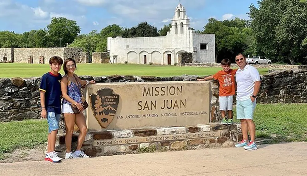 A family of four is posing for a photo next to the Mission San Juan sign at San Antonio Missions National Historical Park