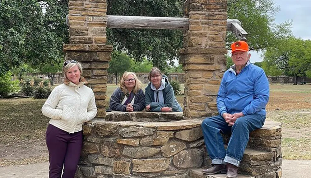 Four people are posing for a photo at an old stone well with three grouped together inside the wells frame and one standing to the side