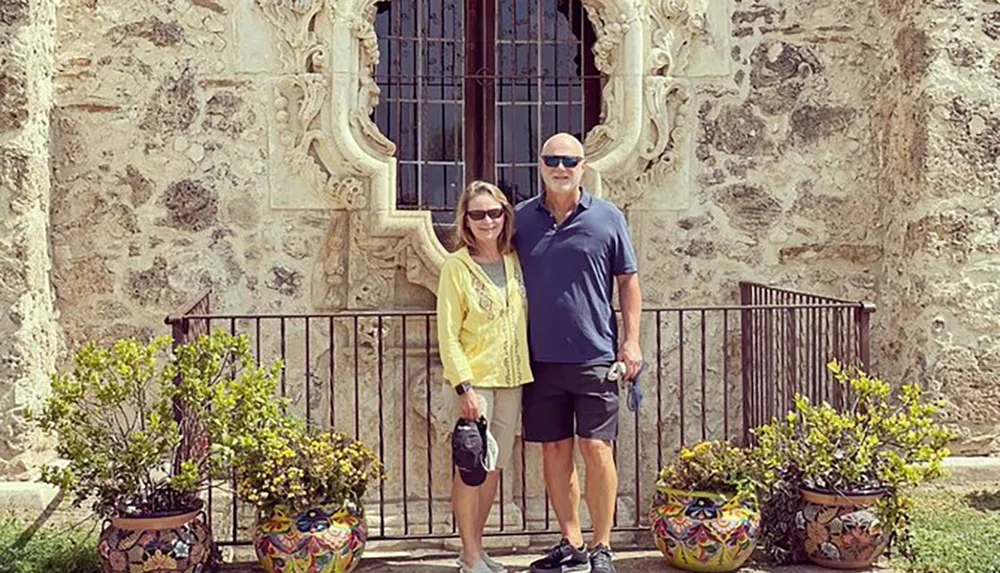 A man and a woman are posing for a photo in front of an ornate historic building with decorative plants around them