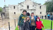 A group of four people are posing for a photo in front of the Alamo on a cloudy day.
