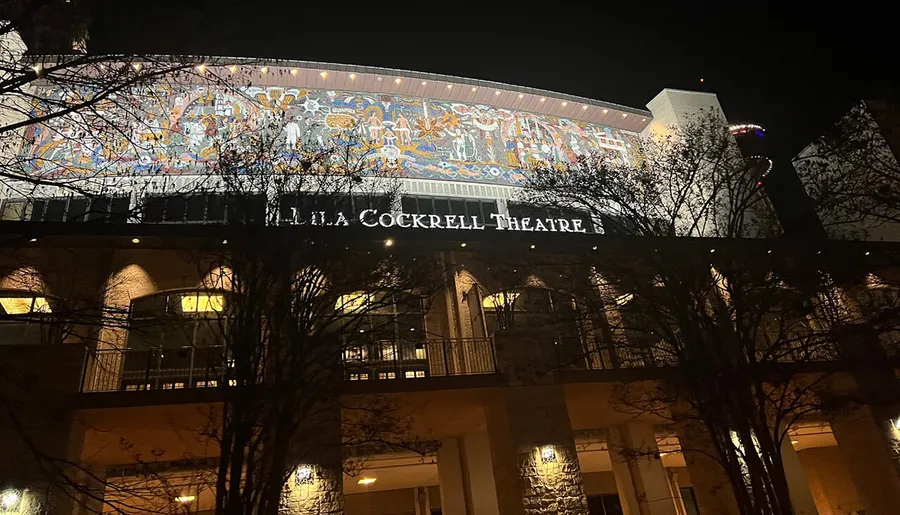The image shows the illuminated exterior of the Lila Cockrell Theatre at night, featuring a large, colorful mural above the entrance sign.