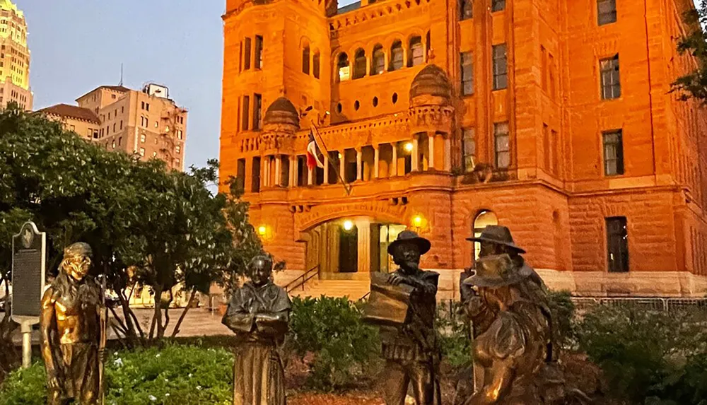 This image features a group of bronze statues in the foreground depicting historical figures with the illuminated ornate facade of a large traditional red sandstone building in the background at twilight