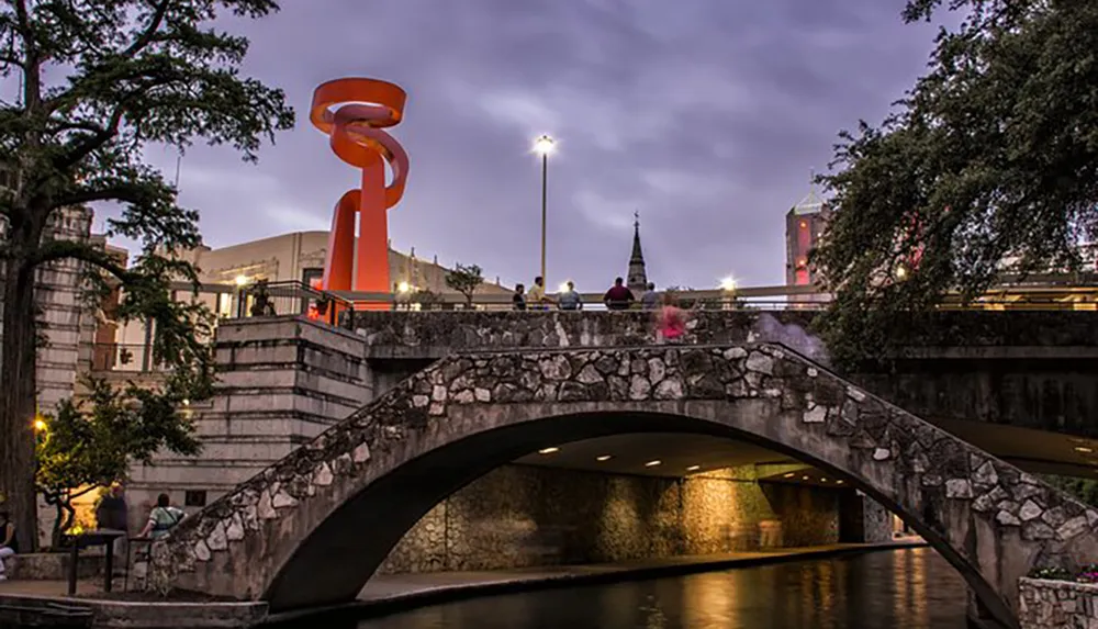 An old stone bridge spans a river in an urban setting illuminated by the glow of streetlights and an orange sculpture at dusk