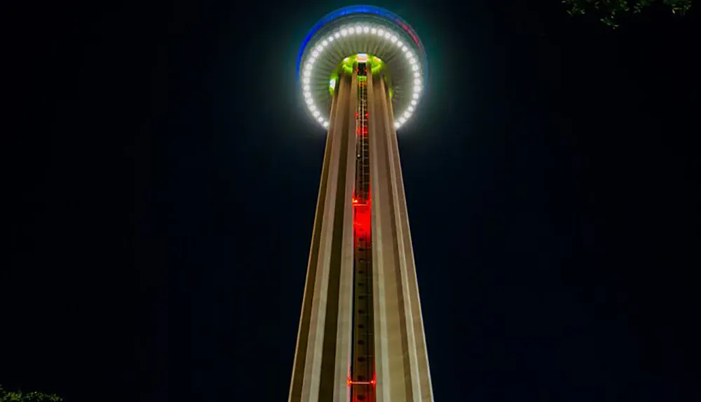 The image shows a tall illuminated tower at night with a circular observation deck at the top glowing with colorful lights against a dark sky