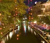 The image shows a vibrant night scene of an illuminated riverwalk with people dining alfresco and a boat gliding on the water