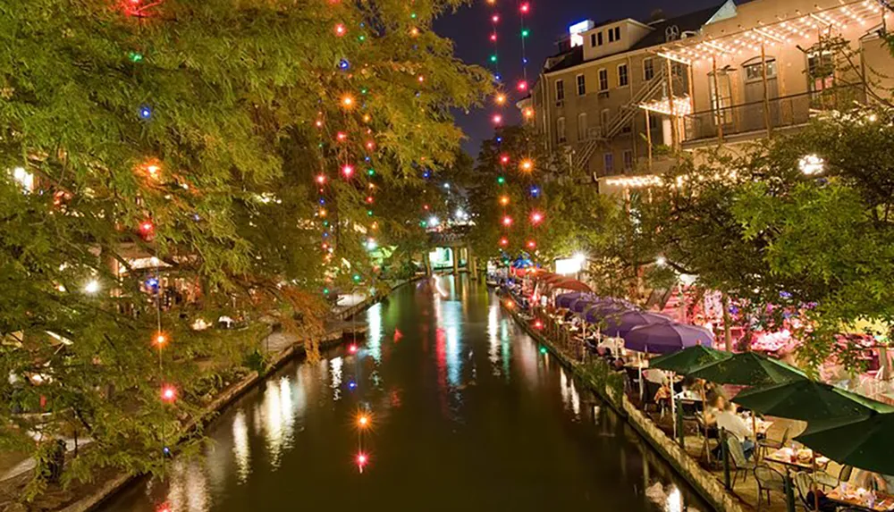 This image shows a vibrant evening scene with colorful lights reflecting on the water of a canal bordered by outdoor restaurant seating under umbrellas and trees adorned with more lights