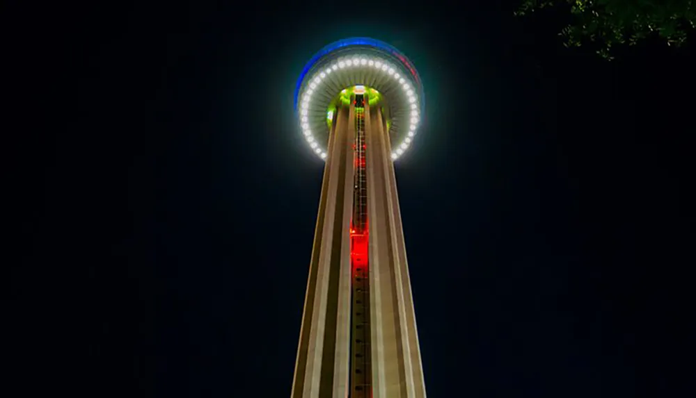 A towering observation tower is illuminated at night with colorful lights highlighting its circular observation deck and long vertical structure