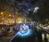 The image shows a vibrant night scene of an illuminated riverwalk with people dining alfresco and a boat gliding on the water