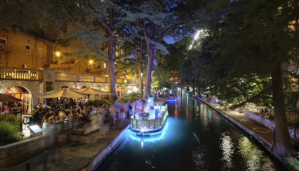 The image shows a vibrant night scene of an illuminated riverwalk with people dining alfresco and a boat gliding on the water