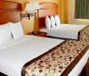 The image displays a tidy and well-lit hotel room with two double beds each adorned with patterned brown runners at the foot a nightstand with a phone a wall-mounted lamp and a window with a green curtain