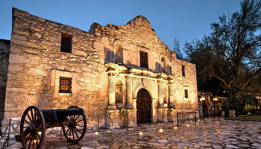 The image shows the historic Alamo mission in San Antonio, Texas, illuminated in the evening.