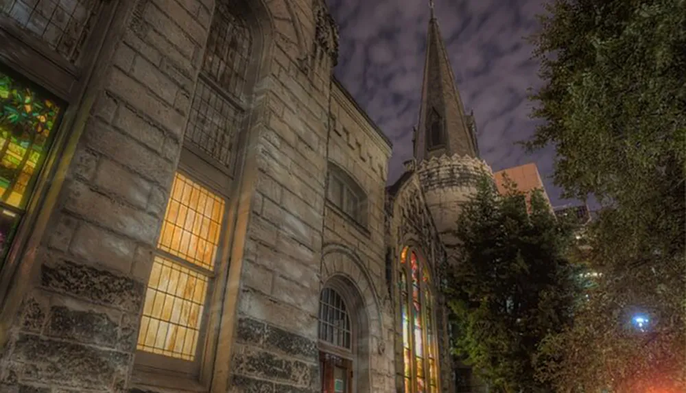 The image shows a historic stone church with stained glass windows illuminated at night under a cloudy sky