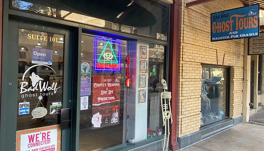 The image shows the exterior of a storefront with the sign Bad Wolf Ghost Tours and Haunted Pub Crawls, alongside a neon sign for Tarot Cards, with various posters in the window and a skeleton decoration standing outside.