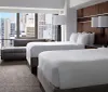 The image shows a modern double hotel room with two beds a sitting area and a large window offering a view of an urban skyline