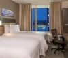 A modern hotel room with two beds features a large window offering a nighttime cityscape view complemented by tasteful decor and a work desk with a television