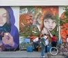 A group of people is seen in front of a vibrant street mural featuring an ethereal figure with a galaxy within her hands juxtaposed with other colorful and fantastical elements