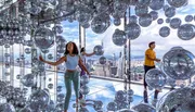 Visitors explore a reflective art installation filled with silvery spheres against the backdrop of a high-rise cityscape seen through large windows.