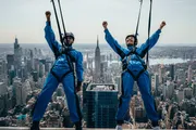 Two people in blue jumpsuits are suspended by safety harnesses high above a cityscape, with one of them raising their arms in excitement.