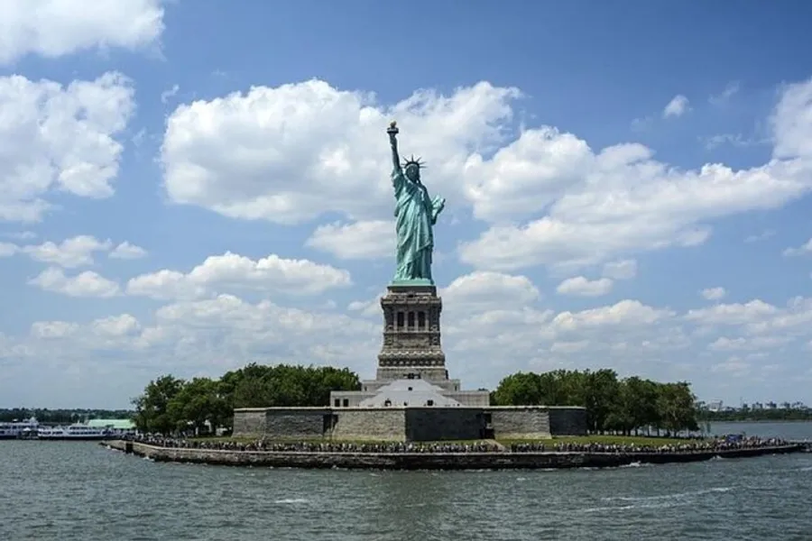 The image shows the Statue of Liberty standing on Liberty Island against a backdrop of blue skies and scattered clouds.