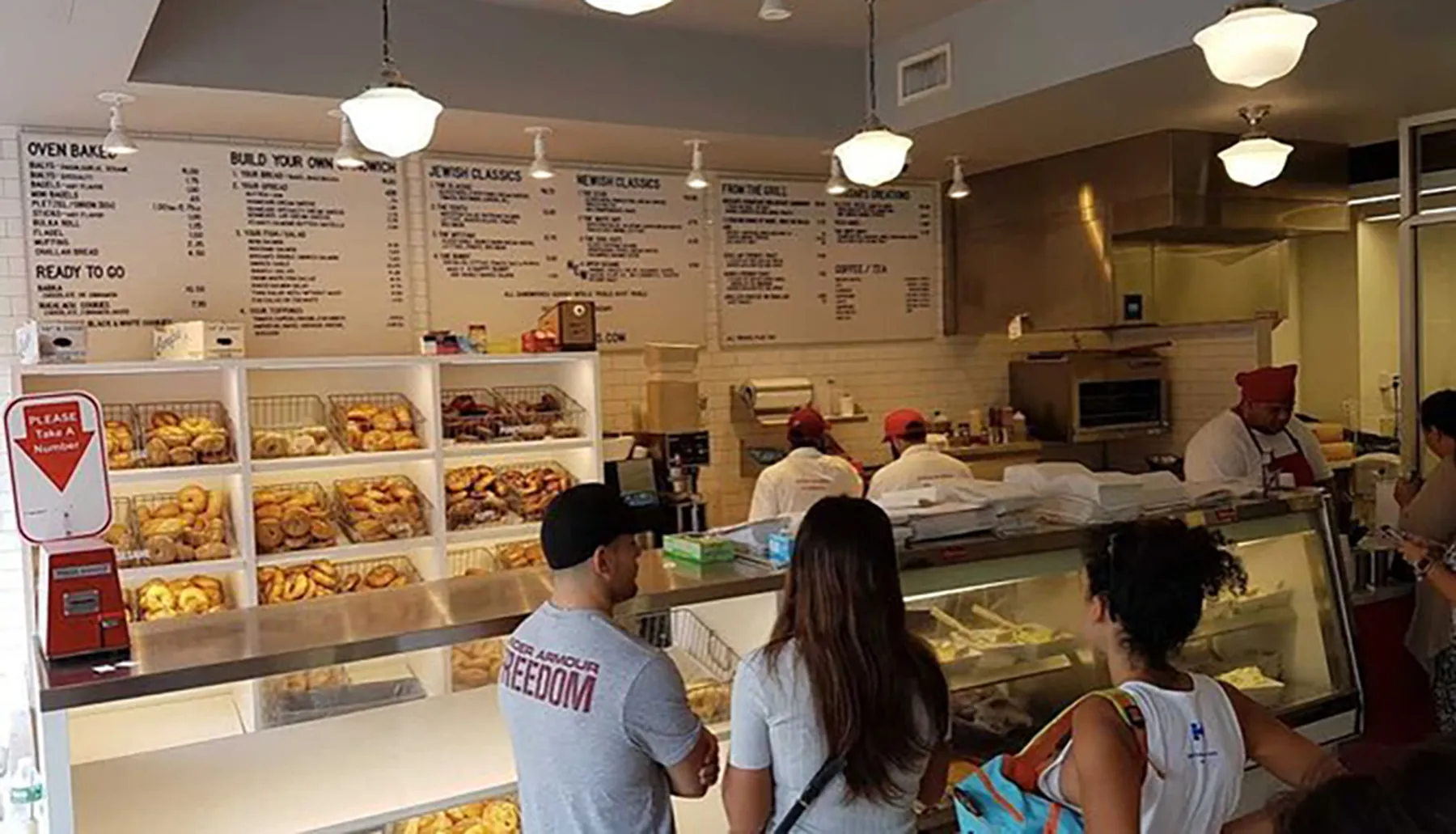 Customers are waiting in line at a deli counter with an array of baked goods on display and a menu board listing various sandwiches and dishes above.