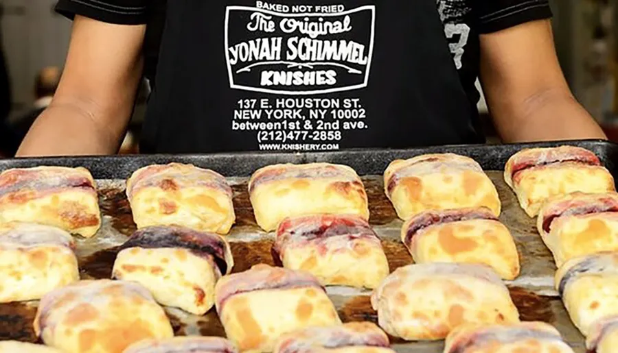 A person is presenting a tray of baked knishes in front of a sign that advertises The Original Yonah Schimmel Knishes in New York City.