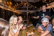 A group of people is enjoying an evening at an outdoor patio, socializing and laughing under string lights.