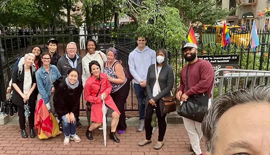 A diverse group of people is posing for a group photo outdoors with some individuals smiling and one person wearing a face mask.