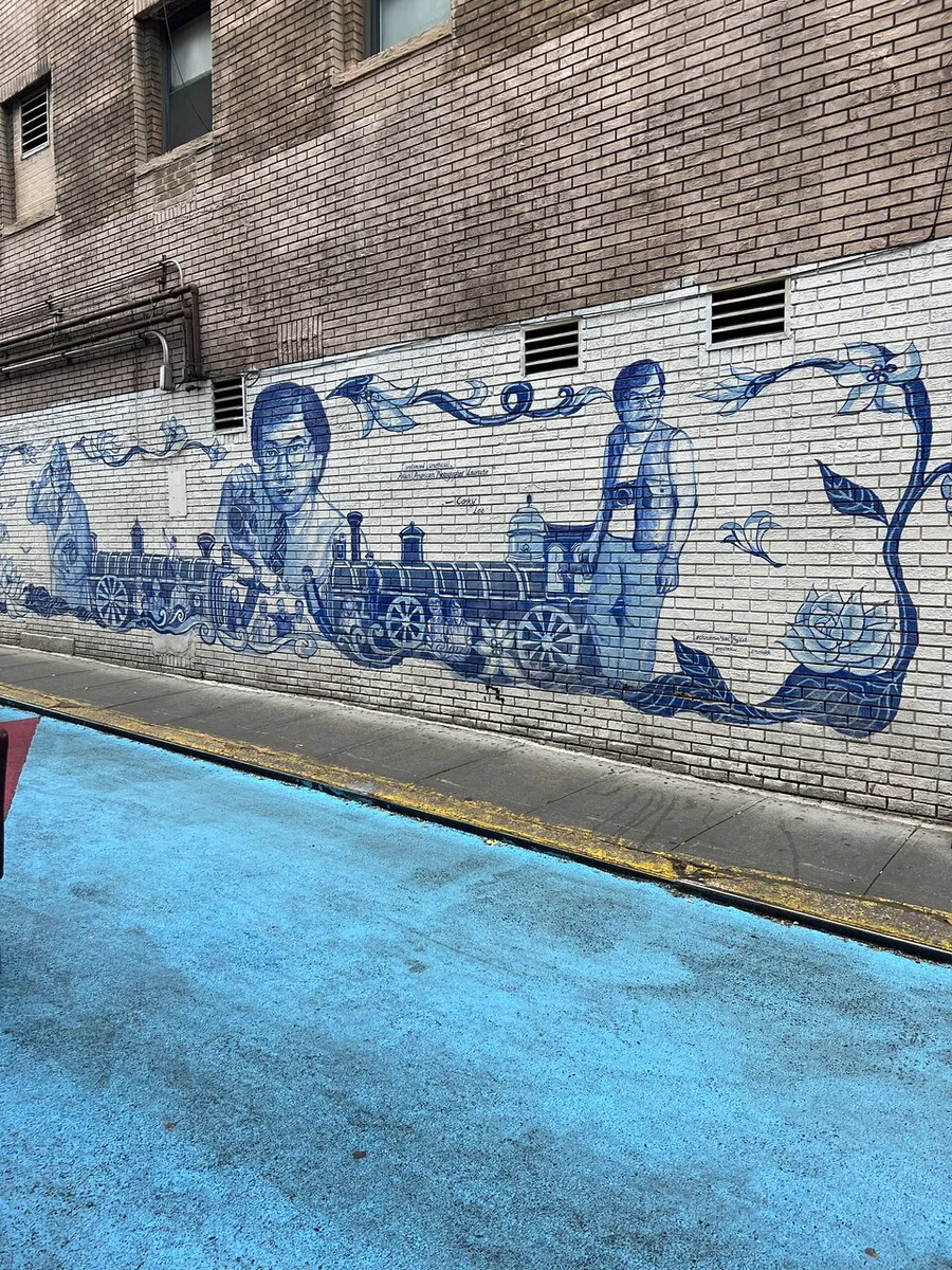 The image shows a detailed blue and white mural on a brick wall depicting individuals and a locomotive, complemented by a similarly colored pavement.