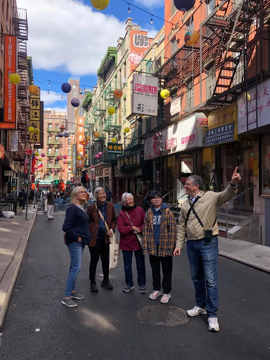 A group of five adults appears to be on a tour, listening to a man pointing out something of interest, in a vibrant street that is likely part of a Chinatown district, flanked by colorful signage and traditional lanterns.
