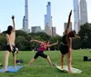 Three people are practicing yoga in a park with skyscrapers in the background