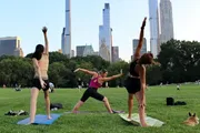 Three people are practicing yoga in a park with skyscrapers in the background.