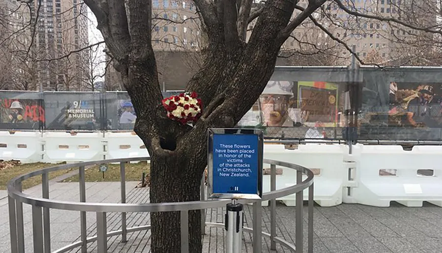 A heart-shaped floral tribute is placed on a tree trunk next to a sign that states the flowers are in honor of the victims of the attacks in Christchurch, New Zealand, with the 9/11 Memorial Museum in the background.