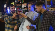 Three men are toasting with glasses of beer in a dimly lit bar setting.