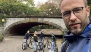 A man takes a selfie in the foreground with four people with bicycles standing under a bridge in the background.