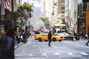A bustling city scene with pedestrians crossing the street, a yellow taxi cab in the foreground, and steam rising from a street vent.